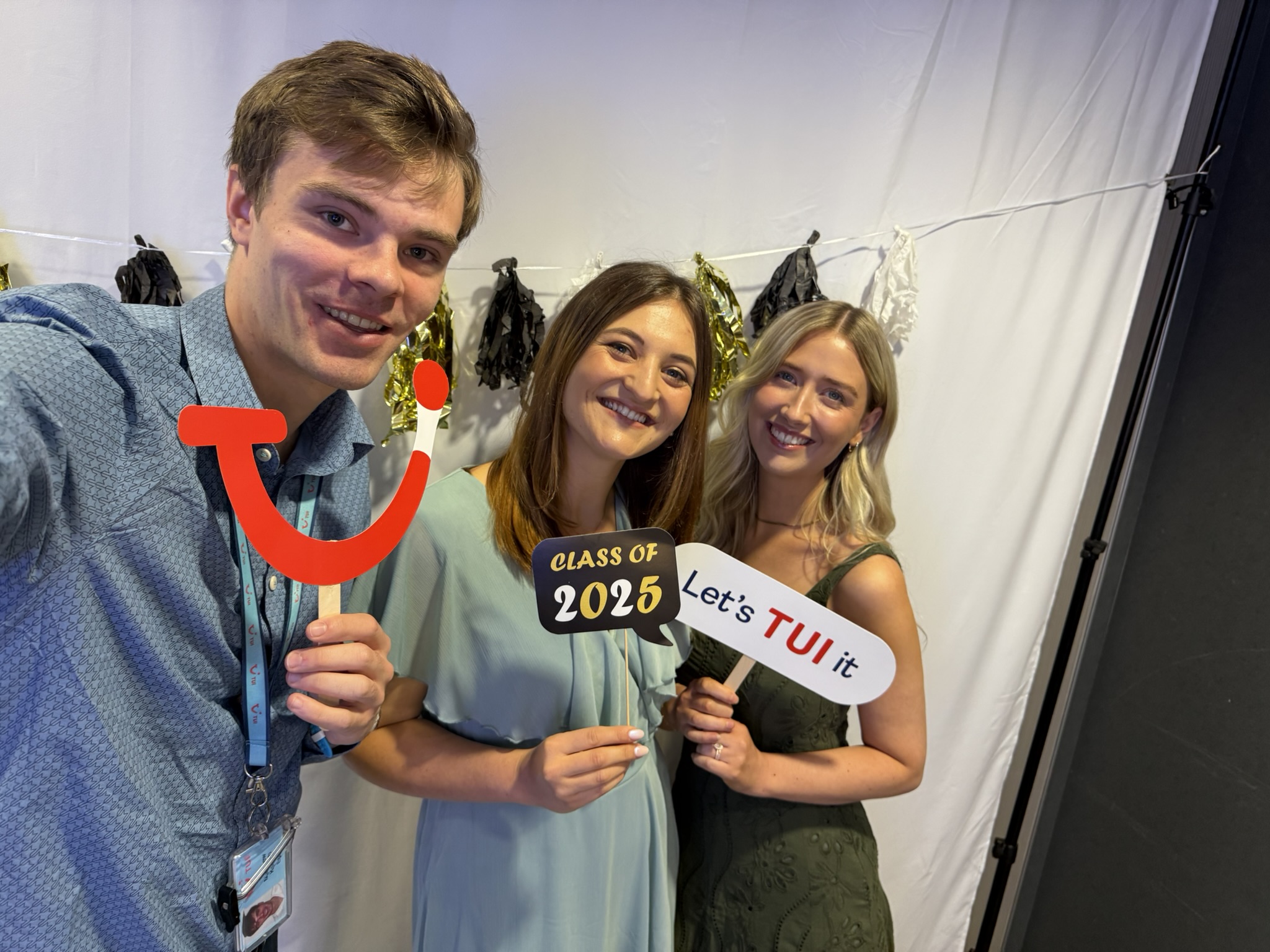 Three smiling people hold props at a celebratory event. One holds a TUI logo, another holds signs saying "Class of 2025" and "Let's TUI it."