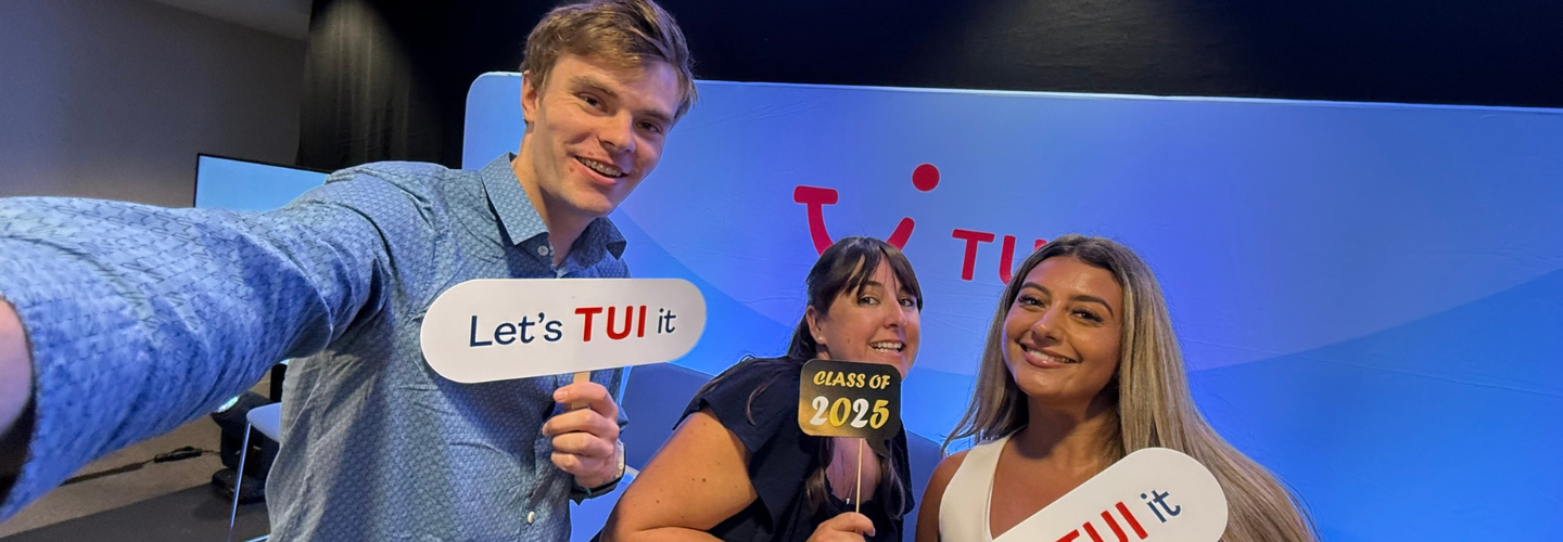 A group of three people smiling taking a selfie, in front of a blue wall with a red TUI logo, holding props that say 'Let's TUI it' and 'class of 2025'.