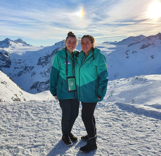 Two Crystal Ski Childcare Reps stand on a mountain smiling, with a snowy background.