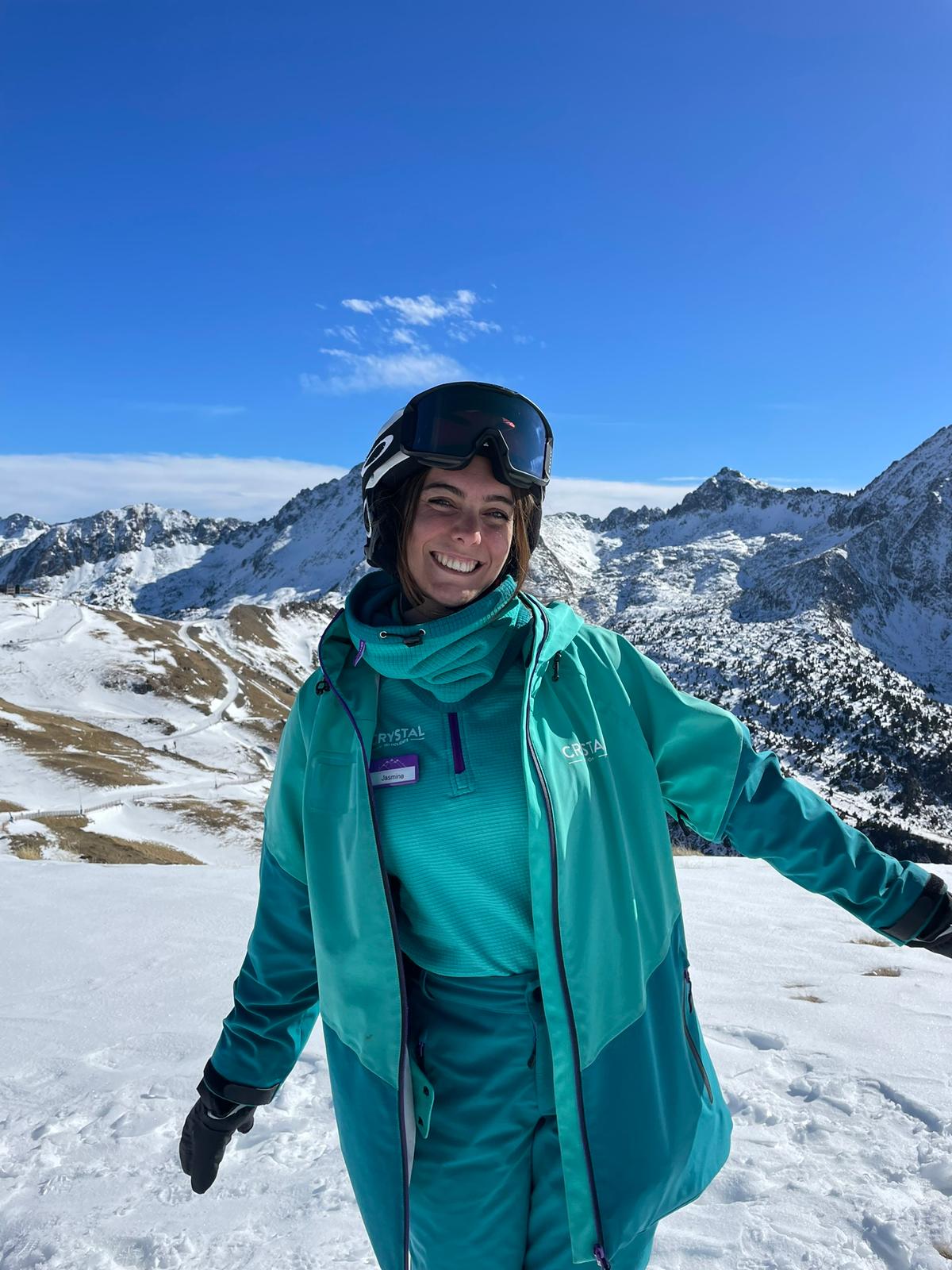 A female Crystal Ski Rep stands on a snowy mountain in Crystal Ski uniform and ski gear.