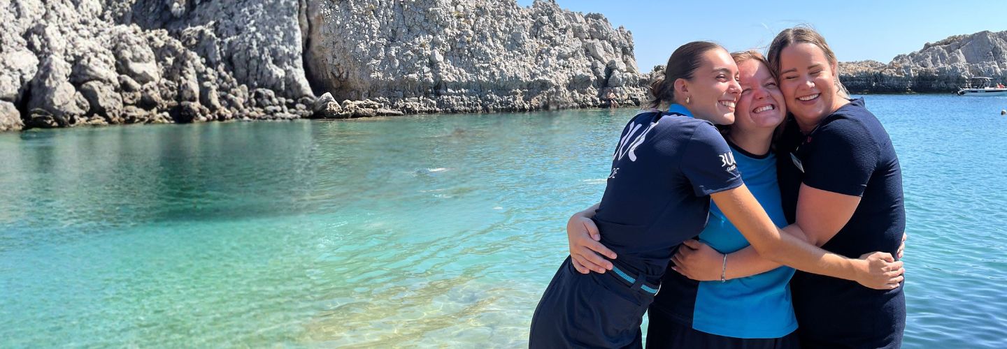 Three female colleagues stand alongside a lagoon in Greece embracing each other