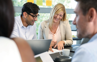 Four colleagues sit at a desk over laptop and paperwork making conversation and laughing.