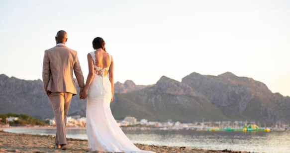 A bride and groom walk holding hands along a beach with a scenic mountain background.