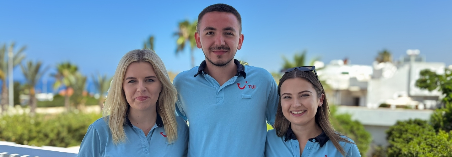 Three TUI Reps in uniform stand smiling together in front of a blue sky and green scenery