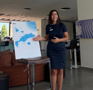 A TUI BLUE GUIDE stands smiling in a hotel next to a poster with a map of Kos