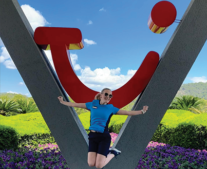 TUI colleague jumps in front of large TUI logo and blue sky.