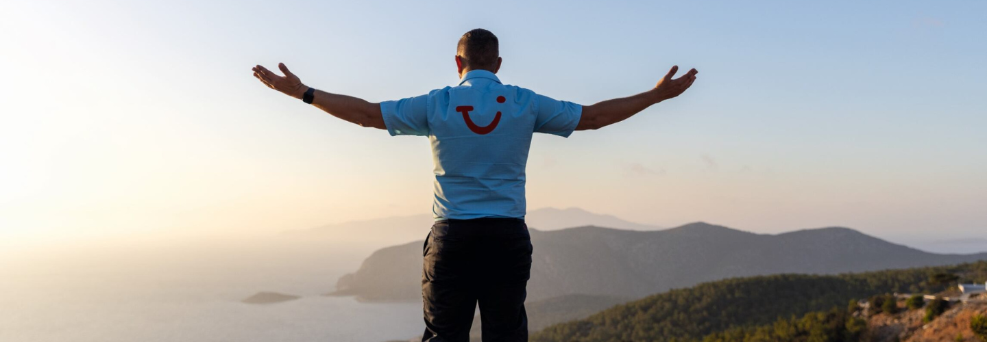 TUI Rep with TUI shirt on faces away from the camera with outstretched arms, to a scenic mountain and sunset.