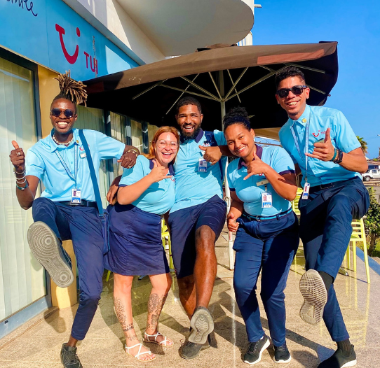 Five TUI male and female TUI Reps stand in front of a TUI office in a fun pose laughing.