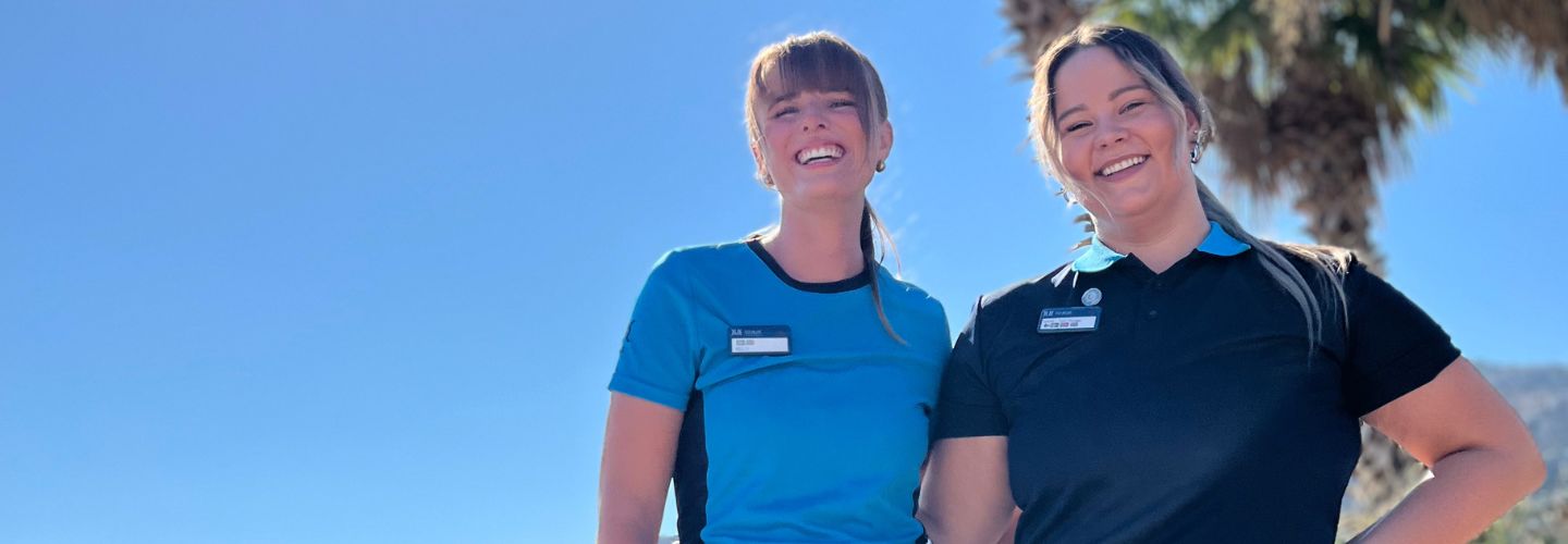 Two TUI BLUE colleagues stand smiling in front of a blue sky and palm trees.