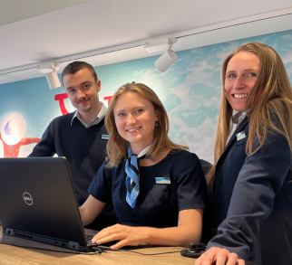 Three travel agents behind the desk with one computer looking and smiling