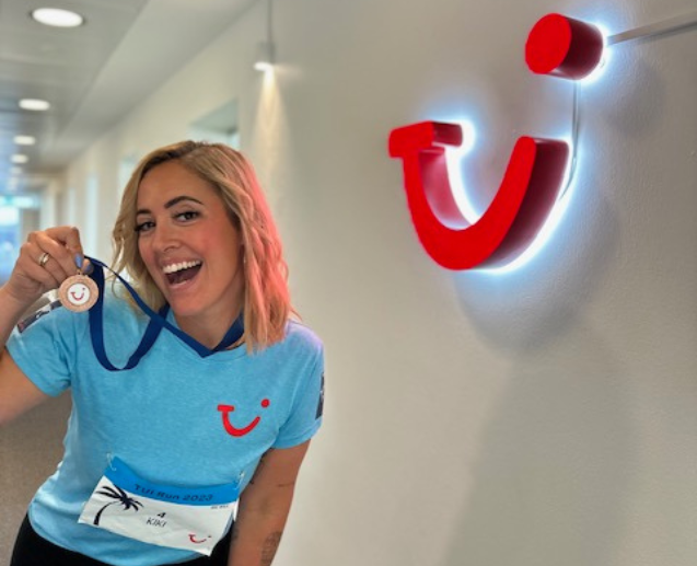 A TUI Denmark female colleague stands in TUI branded sports wear with a medal, in front of a large TUI smile sign.