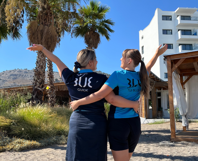 Two TUI BLUE colleagues pose with their back to the camera in front of a Rhodes hotel and palm trees
