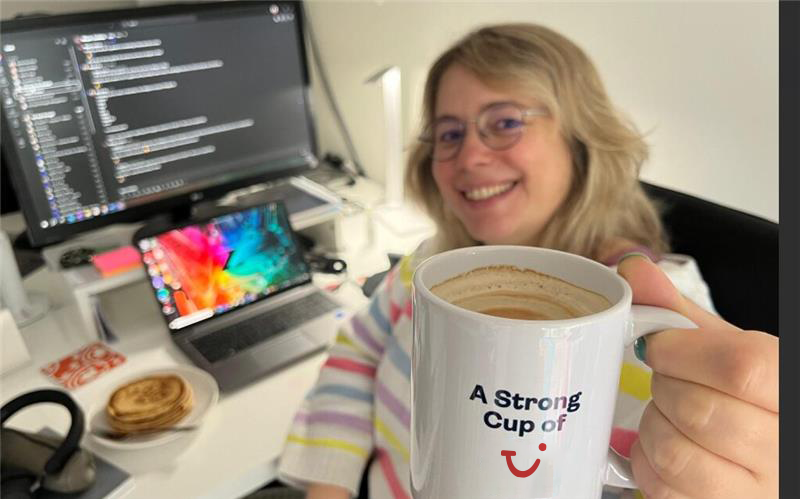 A woman holds a cup with coffee in the background a desk with a laptop and a monitor. 