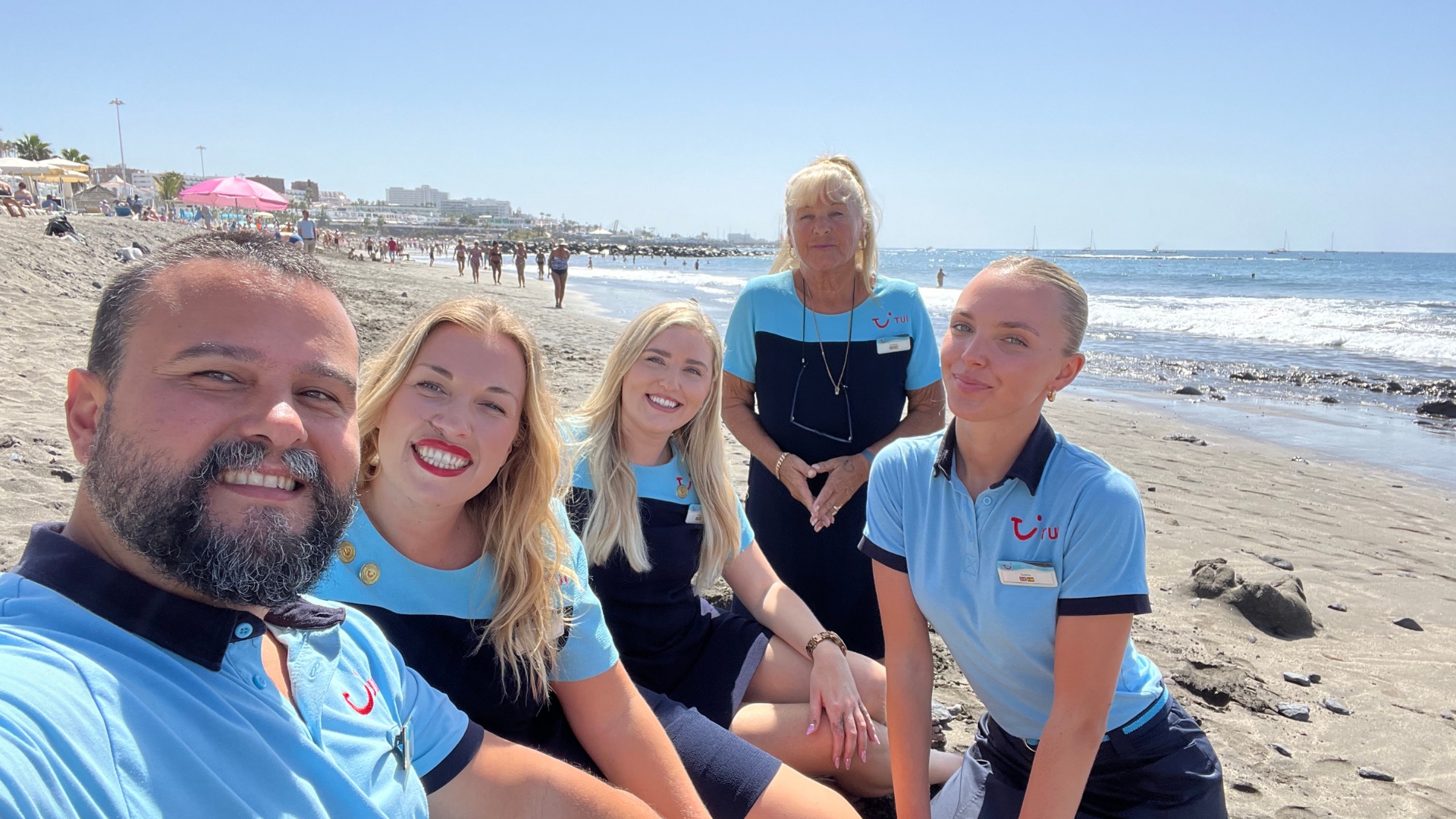 A group of smiling people in TUI Uniform take a selfie on a white sand beach with a blue sky