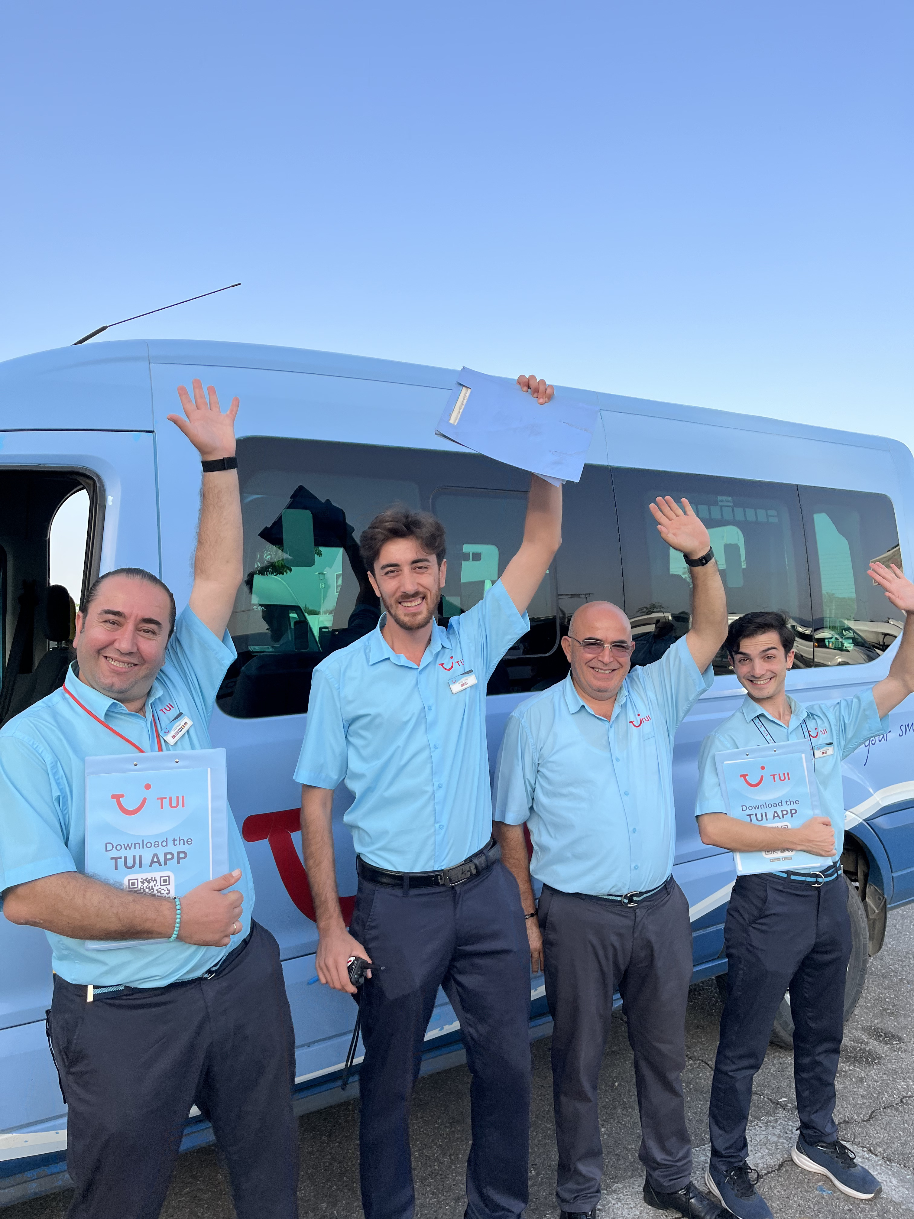 Four TUI Türkiye colleagues stand next to a transfer bus together waving.