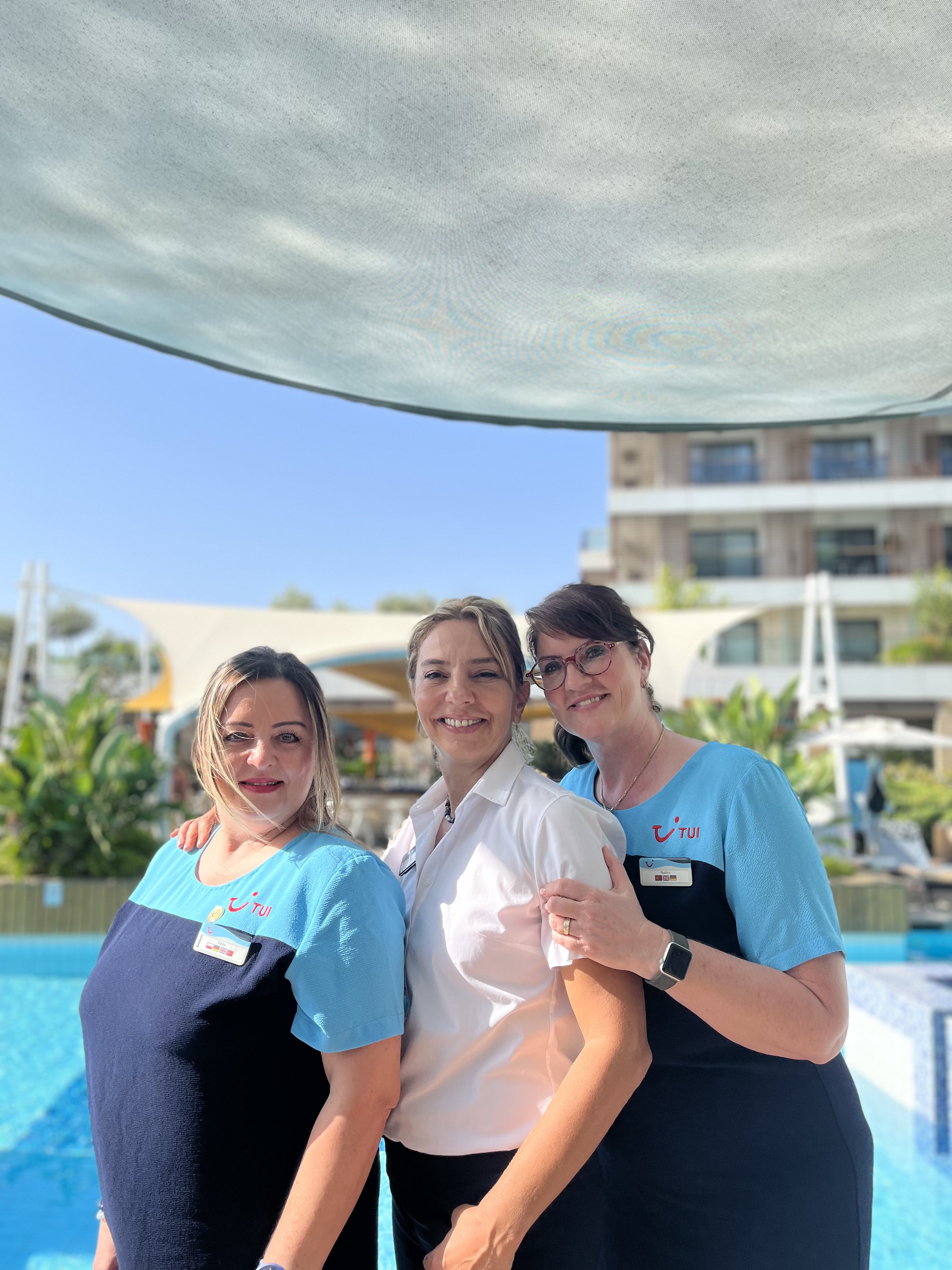 Three female TUI colleagues in front of a pool smiling at the camera