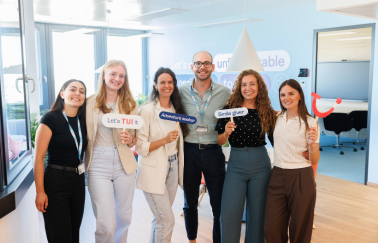 A group of TUI colleagues stand together in a TUI office smiling, holding TUI themed props