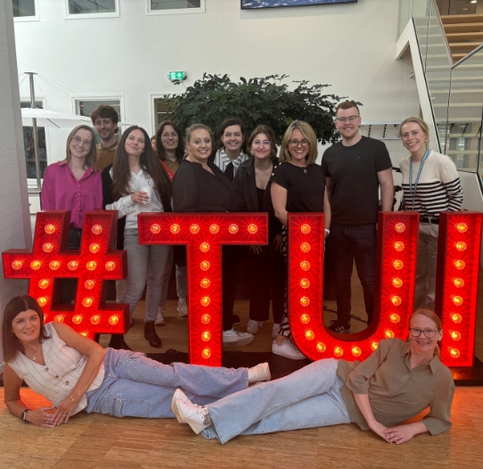 A group of TUI Talent Acquisition colleagues stand together in a TUI office behind a TUI LED sign