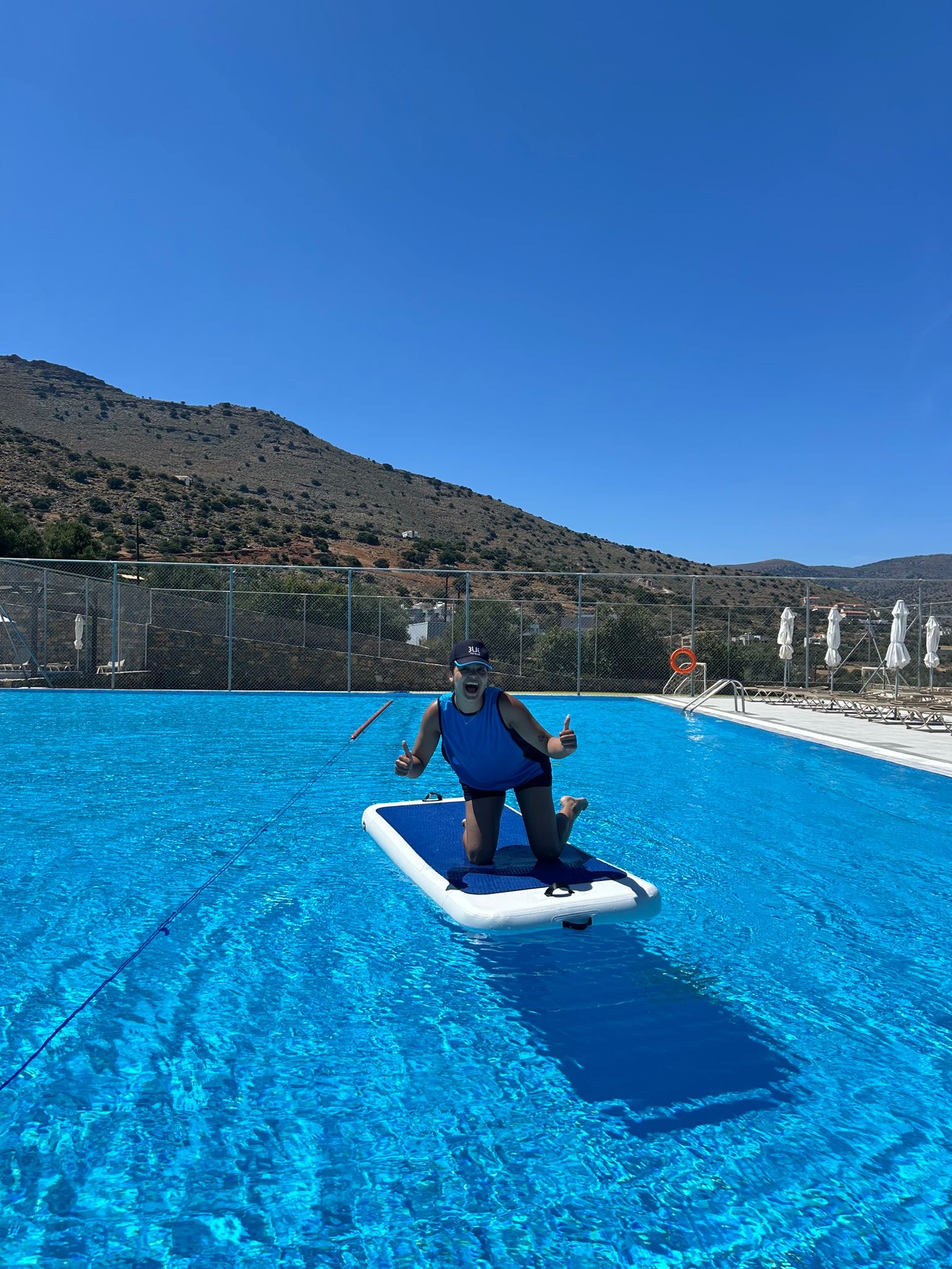 A TUI Fitness Instructor sits on a aqua board placed in a blue pool, with a blue sky behind.