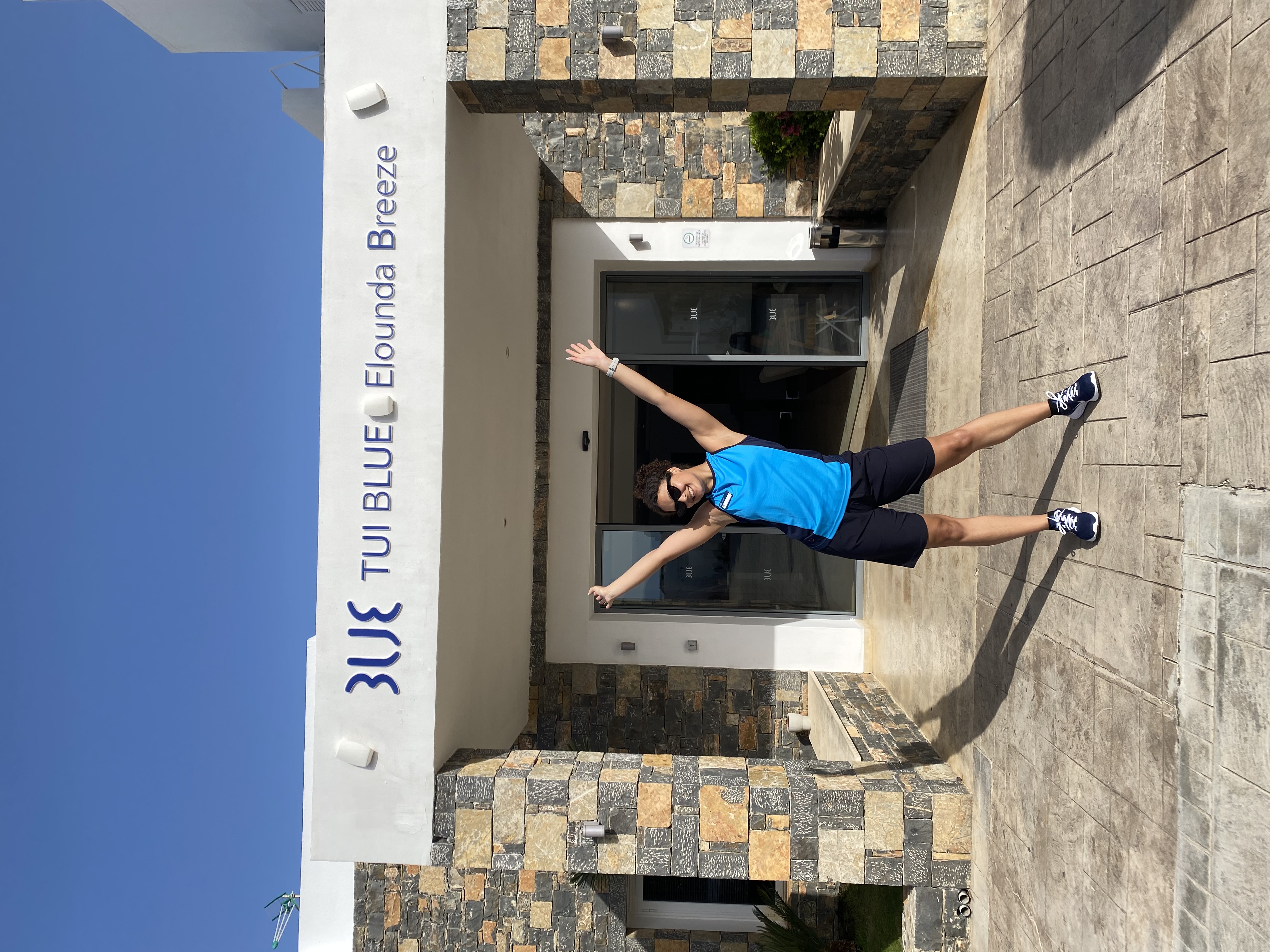 A female TUI BLUE uniformed colleague stands in front of a hotel with her arms in the air.