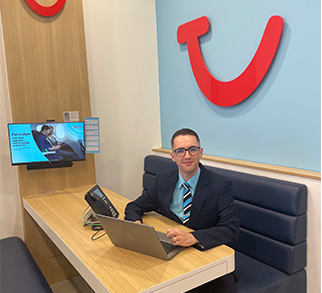 A retail advisor is sat at a booth with the TUI logo behind them. They are looking at the camera, with a laptop on the desk in front of them