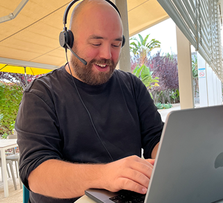 A TUI colleague in a dark jumper sat in front of a MAC laptop outside. They have a headset on with plants in the background on a sunny day. They have a happy expression on their face.