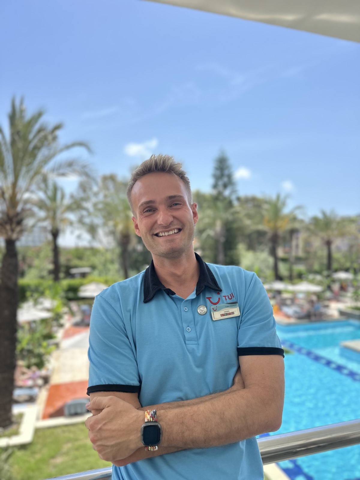 Person in TUI uniform with name tag standing in front of palm trees and a swimming pool.