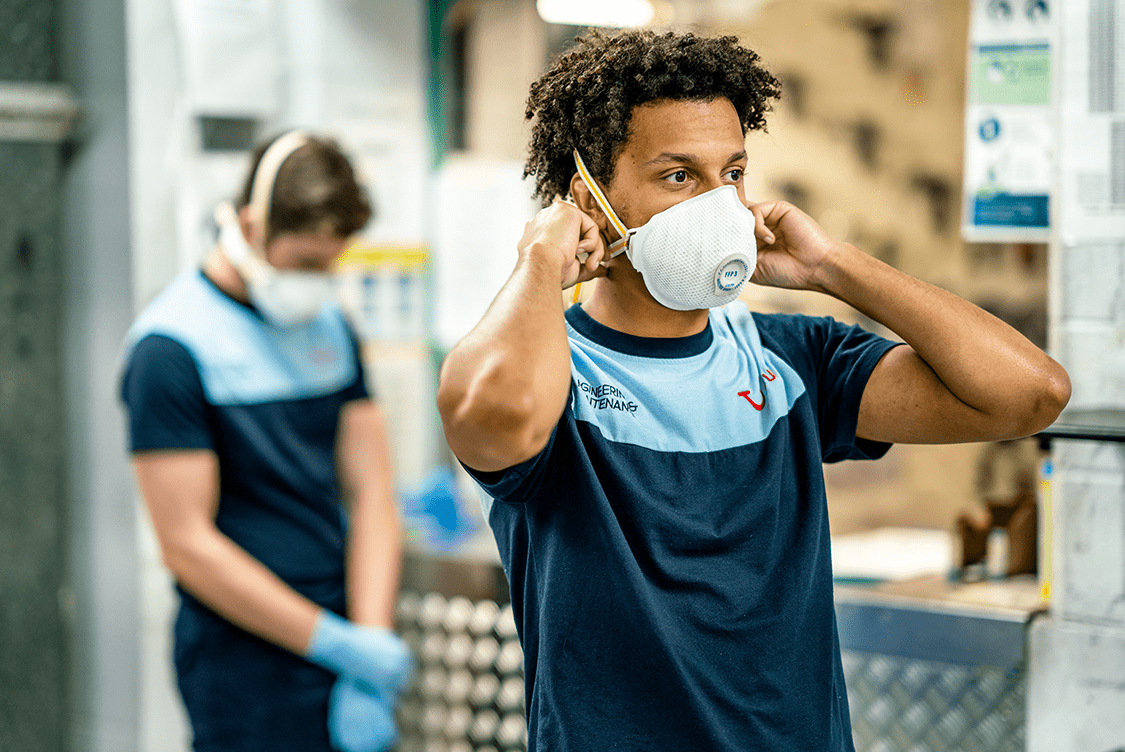 Two workers in a factory wearing blue uniforms and face masks. One adjusts ear protection; the other is focused on a task in the background