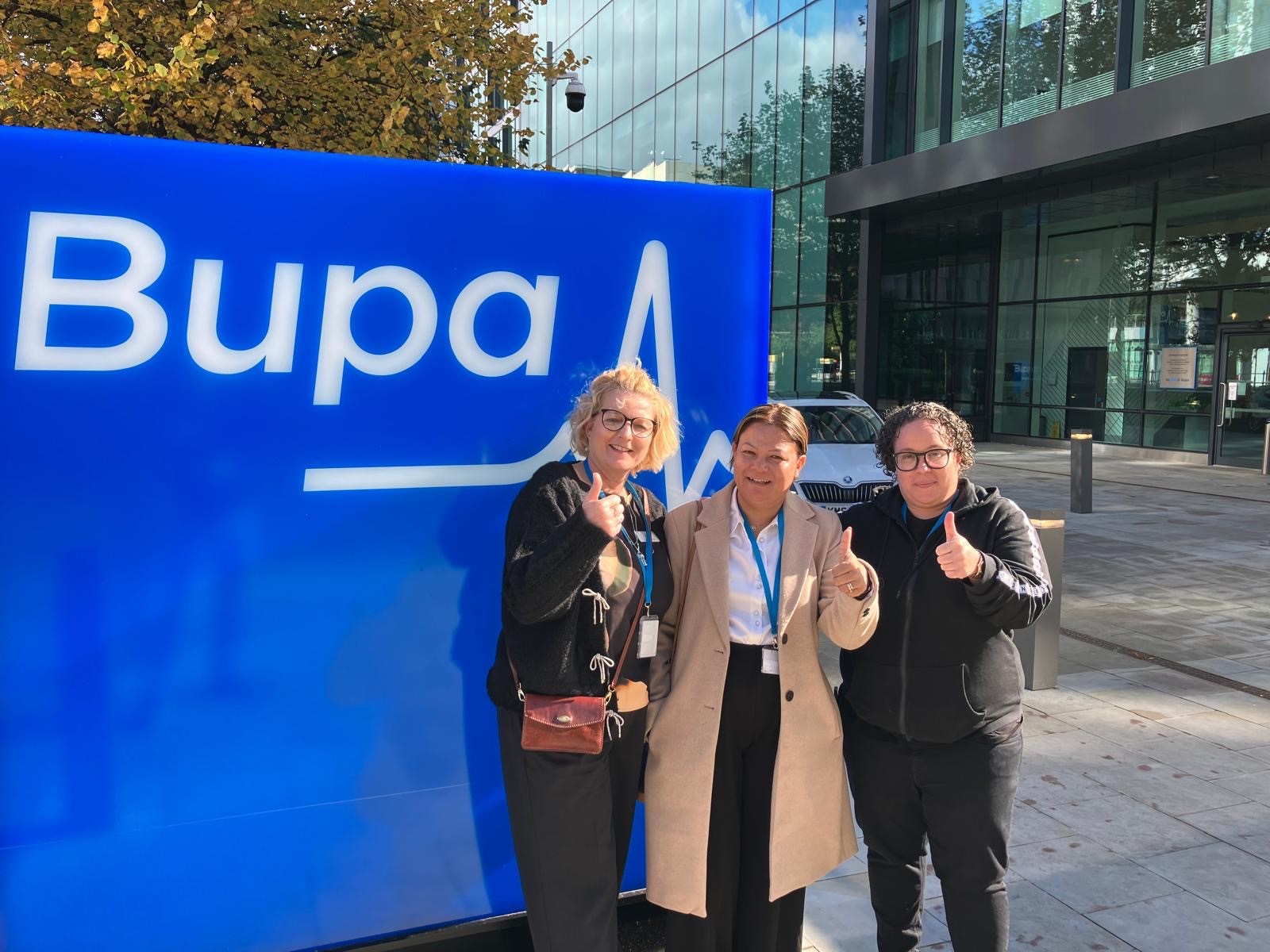 Three people posing thumbs up by Bupa sign