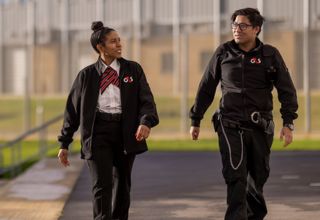 Two people in G4S uniforms walking outside near a fenced area.