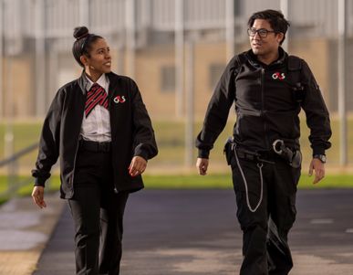Two people in G4S uniforms walking outside near a fenced area.