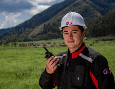 G4S field employee wearing a hard hat and holding a radio in a grassy, rural area.