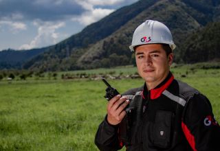G4S field employee wearing a hard hat and holding a radio in a grassy, rural area.