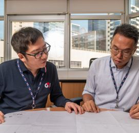Two men reviewing plans at office desk, city view.