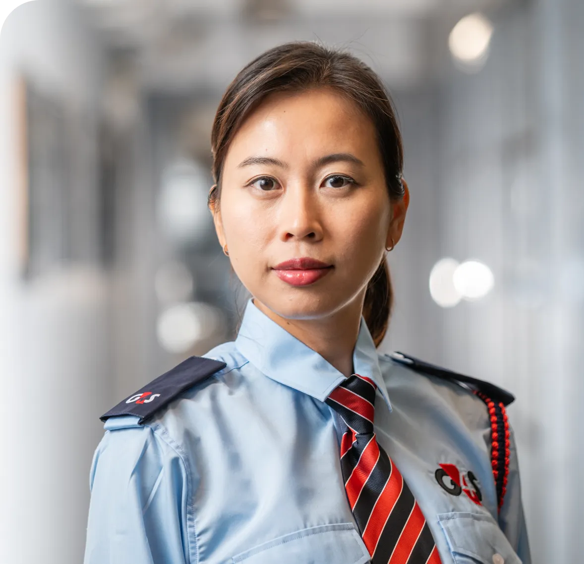 G4S security officer in a light blue uniform with a red and black striped tie stands in a hallway with blurred lights in the background.