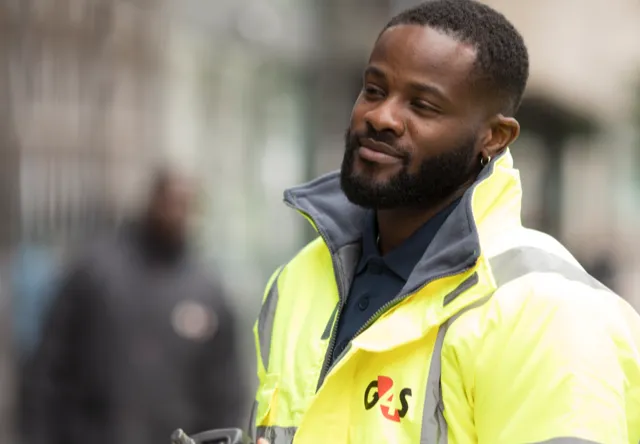 A person in a bright yellow safety jacket stands outdoors, with a blurred urban background hinting at a busy environment.