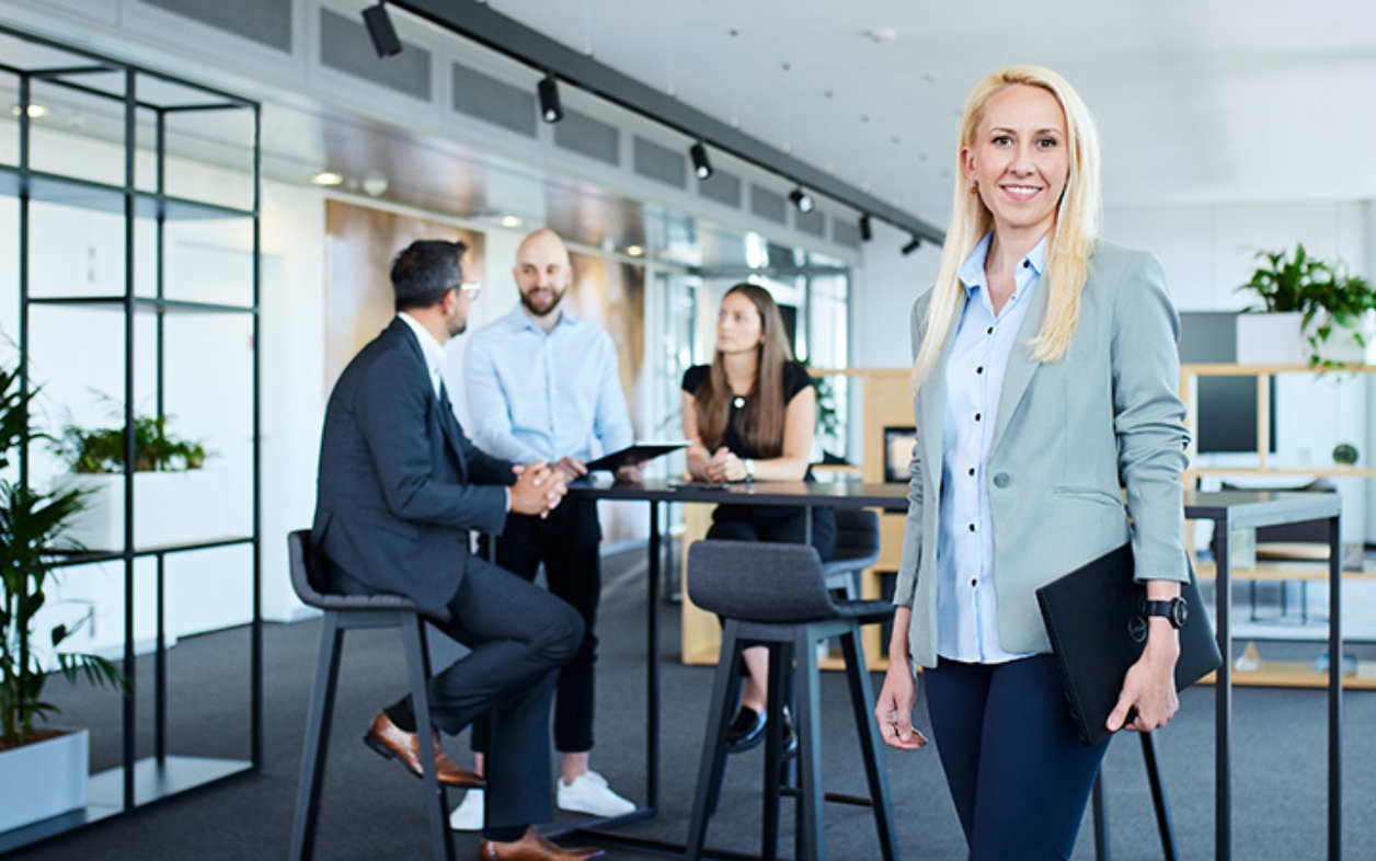 Woman stands smiling in office as team talks behind her