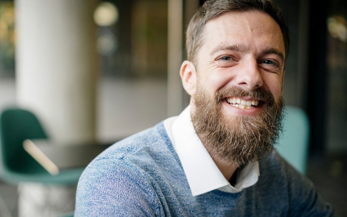 Man in a blue sweater and white shirt, with a beard, smiling into the camera.