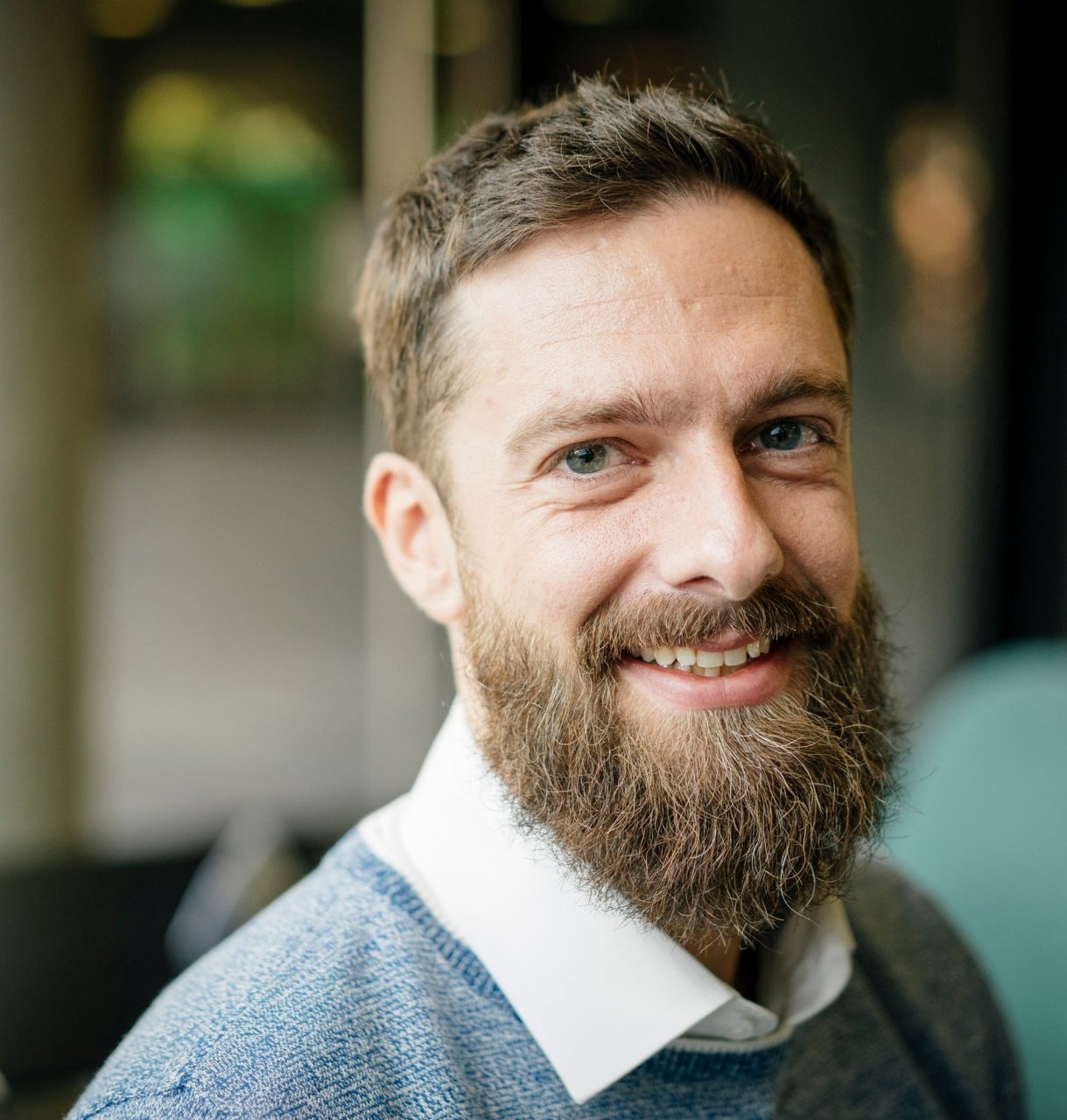 Man in a blue sweater and white shirt, with a beard, smiling into the camera.