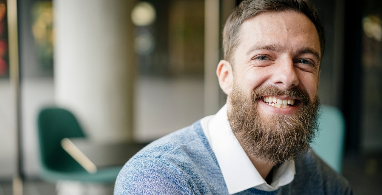Bearded man in a blue sweater with a white shirt underneath smiles at the camera.