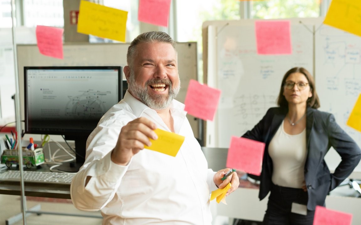 Male colleague working with sticky notes on a screen, woman in the background.