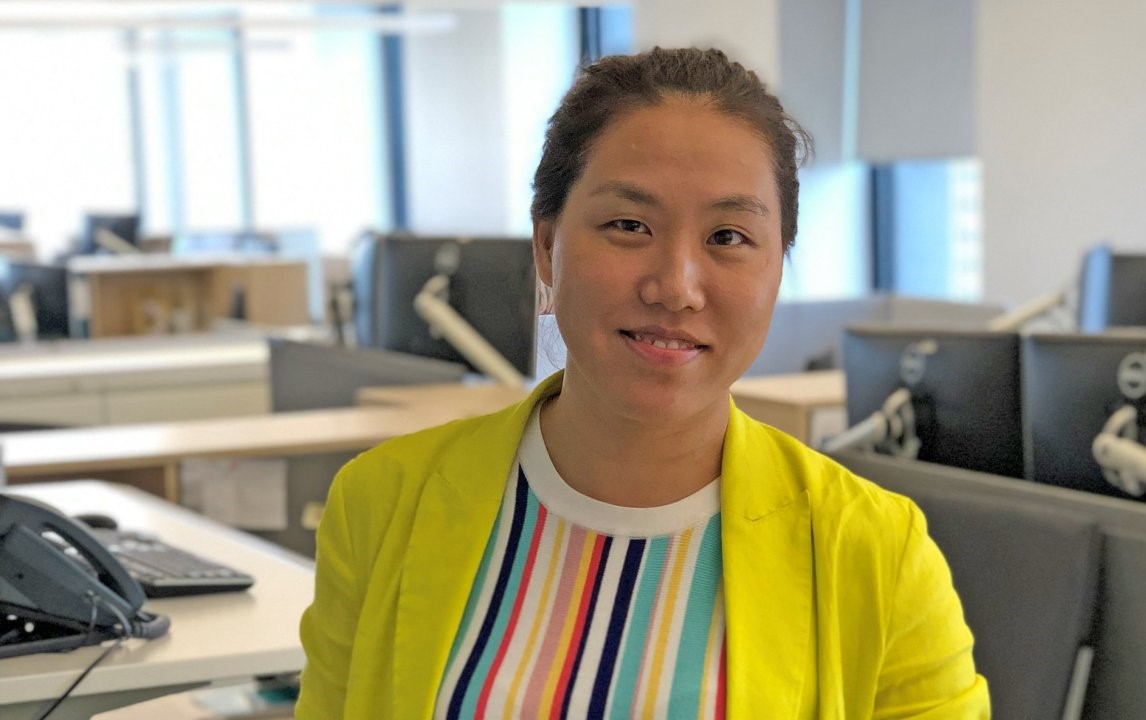 Woman in a colorful shirt in an office environment, smiling into the camera.
