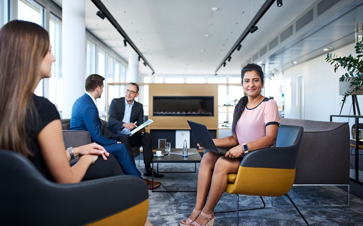 People sitting in a modern office lounge, with one woman holding a laptop and smiling at the camera.