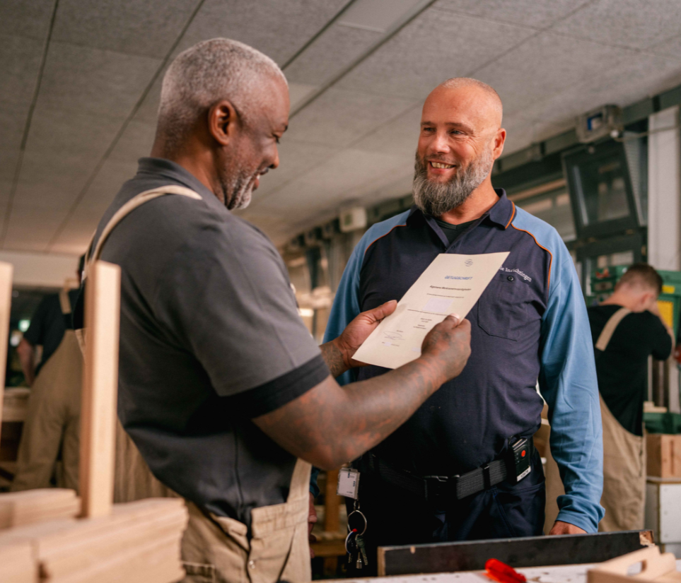 twee mannen naast elkaar. Een man in uniform van DJI en de ander met werkkleding. Ze staan naast een timmerbank.