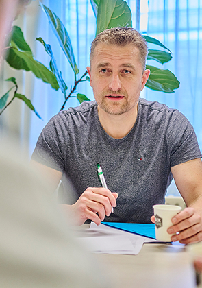 Man met baard in grijs T-shirt praat serieus tijdens overleg aan tafel met pen en koffie