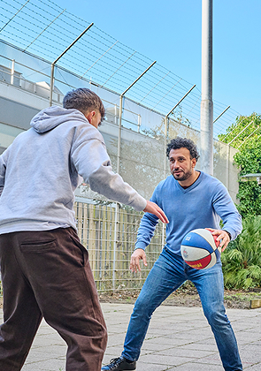 Twee mannen spelen basketbal buiten, twee vrouwen kijken toe op stoelen