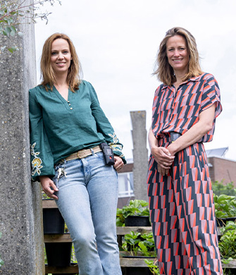 Twee vrouwen staan lachend in een tuin bij plantenbakken
