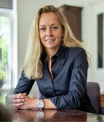 een vrouw met blond haar, zit aan een bureau en kijkt in de camera.