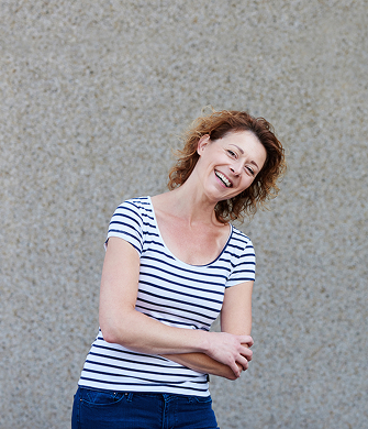Vrouw staat voor een grijze muur. Ze draagt een t-shirt met blauw, witte strepen. Ze kijkt vrolijk in de camera.