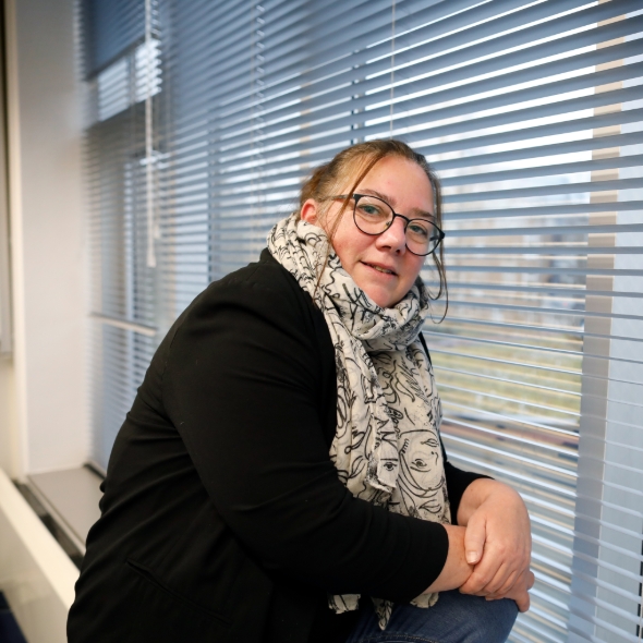 Jonge vrouw met bril en sjaal zit bij raam met luxaflex